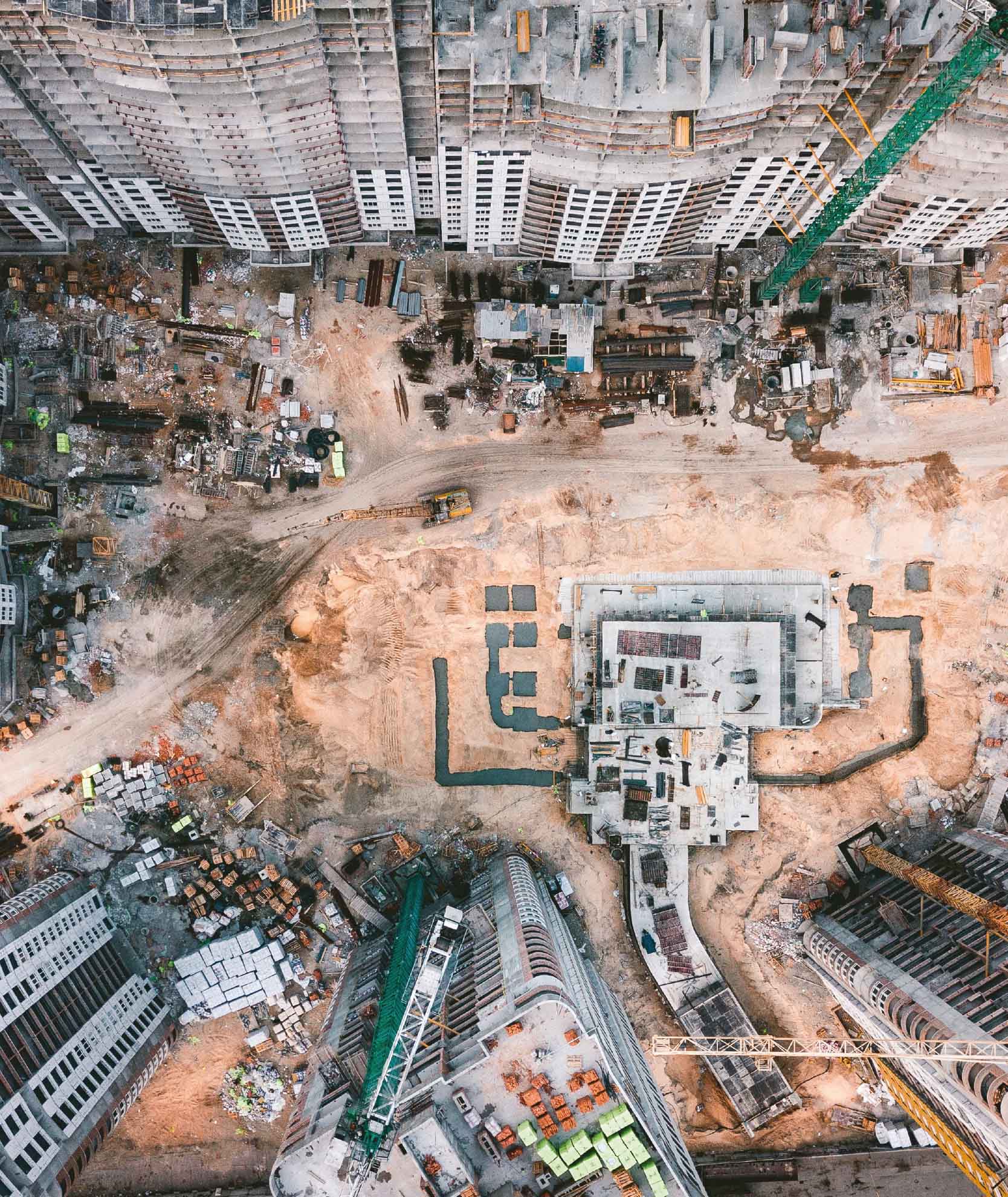 Overhead view of a large construction site with multiple buildings under development, heavy machinery in operation, and various materials spread across the site. Represents comprehensive CDM (Construction Design and Management) support ensuring safe and compliant construction practices