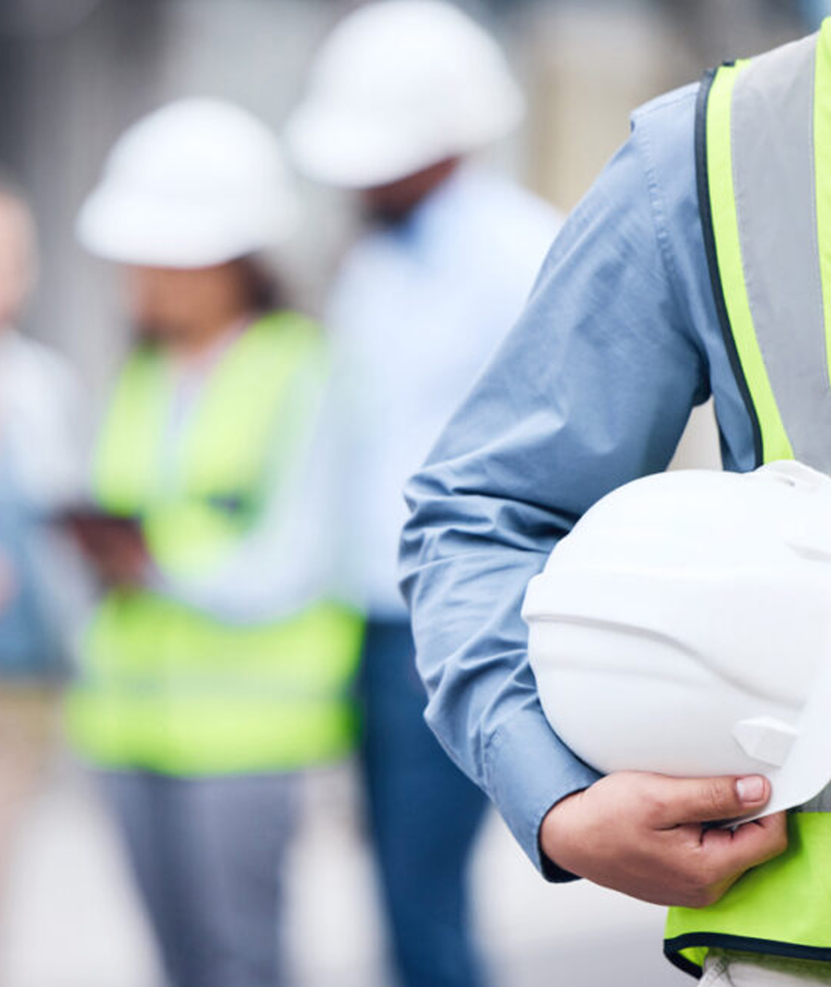Close-up of a professional holding a hard hat, with safety personnel in the background, representing health, safety, and environmental consultancy services for construction, manufacturing, recycling, and logistics industries.